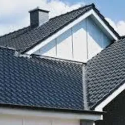 Ciemna dachówka ceramiczna na jasnym domu jednorodzinnym, widok z dołu, fragment rynny, komin w tle, błękitne niebo.