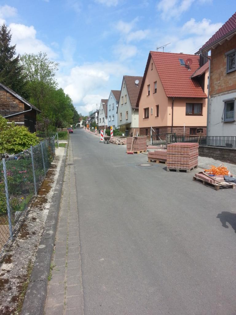 Przygotowane palety z czerwoną kostką brukową na ulicy w zabudowie jednorodzinnej, czekające na ułożenie, widoczne zabezpieczenia drogowe i narzędzia.