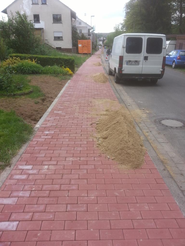 Czerwona kostka brukowa ułożona na chodniku, z widocznymi stertami piasku przygotowanego do spoinowania, biały samochód dostawczy zaparkowany obok.