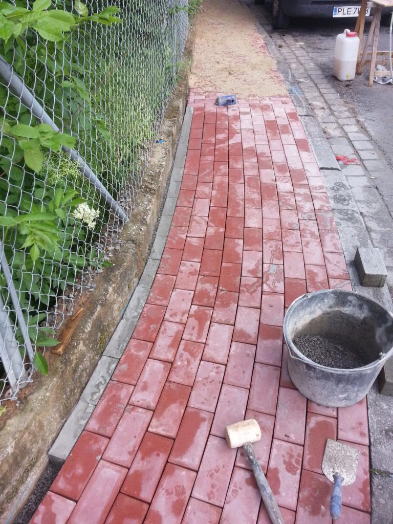 Układanie czerwonej kostki brukowej przy ogrodzeniu z siatki, widoczny gumowy młotek, kielnia i wiadro z zaprawą, częściowo podsypana piaskiem