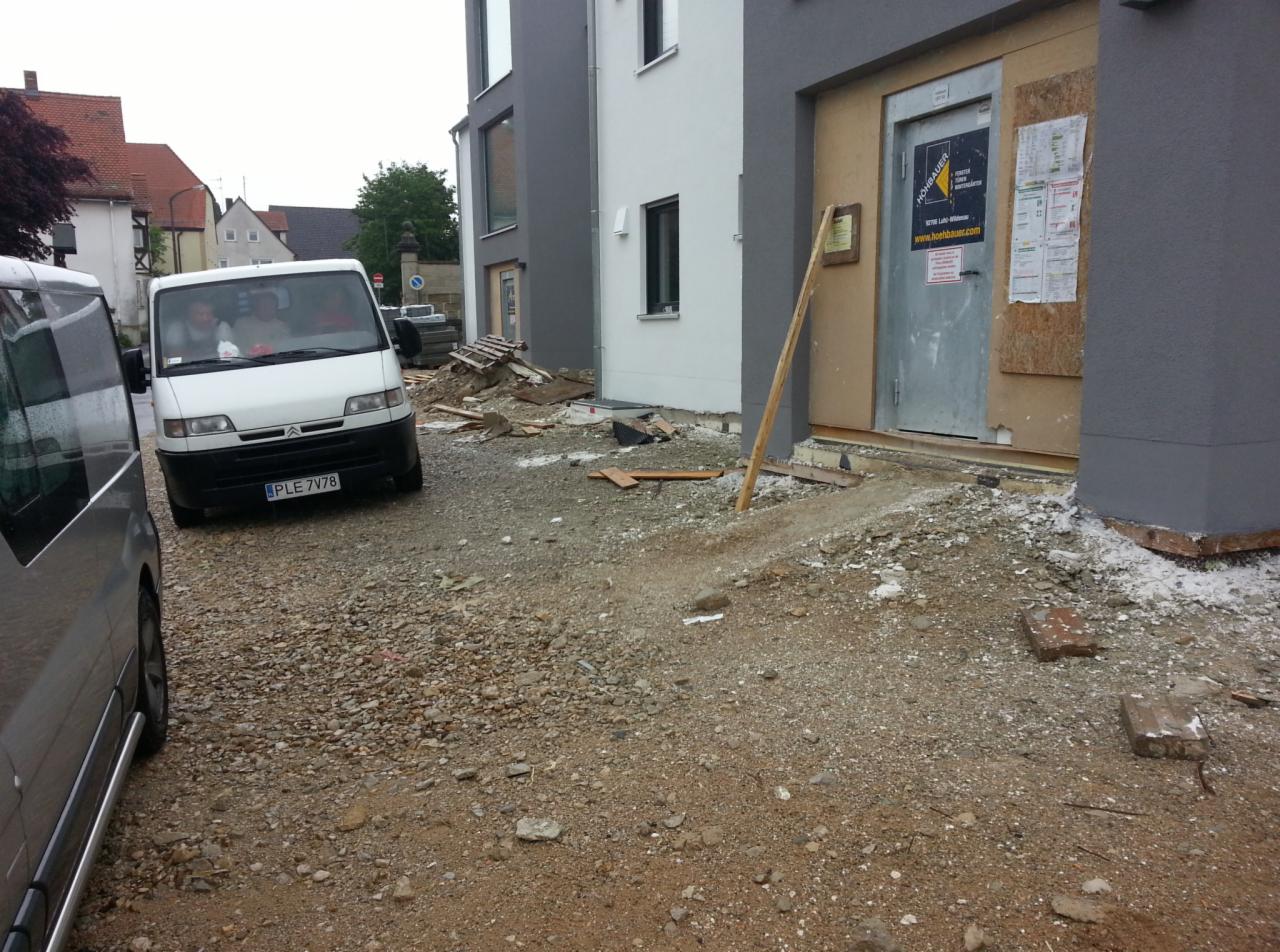 Przygotowanie terenu pod budynkiem: gruz, piasek, częściowo ułożona ścieżka z betonu, zaparkowany biały bus z rejestracją PLE 7V78 i szary samochód osobowy w tle.