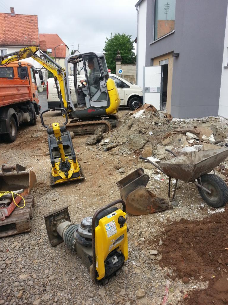Żółty mini-koparka i zagęszczarki na placu budowy przed budynkiem z szarą elewacją, w tle pomarańczowa wywrotka i biały van.