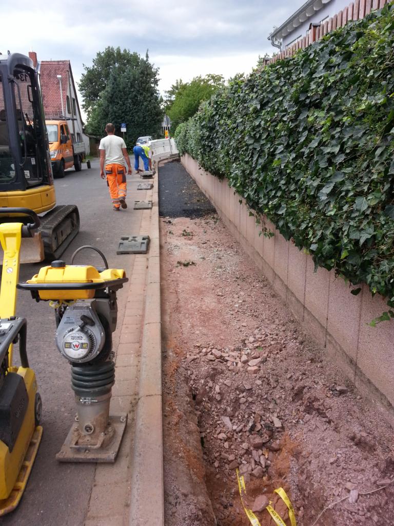 Przygotowanie podłoża pod chodnik z wykorzystaniem ubijarki wibracyjnej; w tle pracownicy w odblaskowych ubraniach oraz mała koparka.