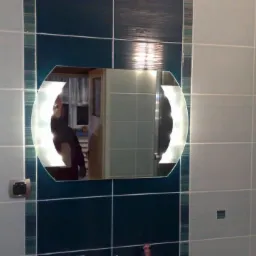 Biało-turkusowa łazienka z podświetlanym lustrem o nietypowym kształcie, otoczonym dekoracyjnymi paskami. Widoczne płytki ceramiczne na ścianach i odbicie osoby w lustrze.