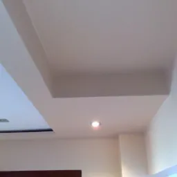 Wykończony sufit podwieszany z oświetleniem punktowym, widoczny fragment ściany i drzwi. Gładka, jasna powierzchnia. Minimalistyczny design.