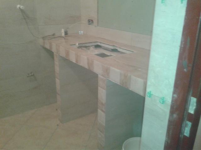 Wykończona płytkami zabudowa pod umywalkę w łazience w trakcie remontu. Widoczne krzyżyki dystansowe, szary odcień płytek, surowy stan wykończenia ścian.