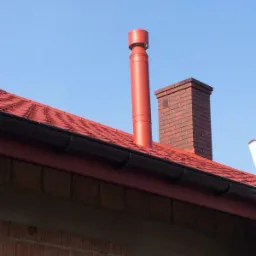Czerwony komin systemowy i komin z cegły na dachu z czerwoną dachówką, widok z dołu na tle błękitnego nieba.