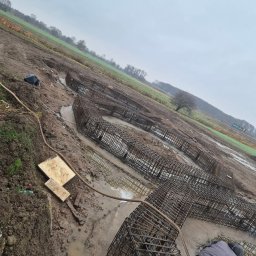 H&R PROJEKT SP. ZO.O. - Zbrojenie fundamentów w wykopie wypełnionym wodą, widoczne drewniane deski i pracownik z narzędziem, pochmurne niebo i pole w tle.