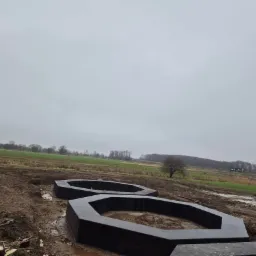 Dwa betonowe, heksagonalne pierścienie fundamentowe w grząskim terenie pod budowę, widok na pole w pochmurny dzień.