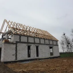 Budowa domu z białych bloczków: surowe ściany z otworami okiennymi i drewniana konstrukcja dachu na tle pochmurnego nieba i drzew.