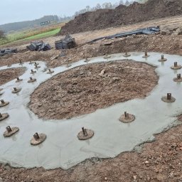 Wylany fundament pod budowę z widocznymi metalowymi elementami mocującymi. Na drugim planie ziemia, trawa i drzewa. Pochmurny dzień.