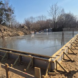 H&R PROJEKT SP. ZO.O. - Zalany fundament pod budowę domu, z drewnianym szalunkiem i wystającymi prętami zbrojeniowymi. W tle widoczne drzewa i ziemia.