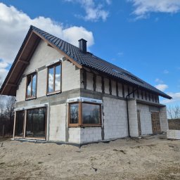 H&R PROJEKT SP. ZO.O. - Surowy stan domu z bloczków, z oknami i dachem, na piaszczystym terenie. Widoczne materiały budowlane. Niebieskie niebo z chmurami w tle. Dom w trakcie budowy.