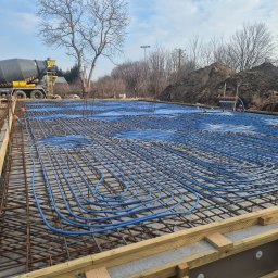 H&R PROJEKT SP. ZO.O. - Fundament z ułożonym zbrojeniem i niebieskimi rurami ogrzewania podłogowego, przygotowany do zalania betonem. W tle betoniarka i hałda ziemi.