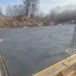 H&R PROJEKT SP. ZO.O. - Zalany fundament pod budowę domu jednorodzinnego, widoczne szalunki i wystające pręty zbrojeniowe. W tle hałda ziemi i drzewa.