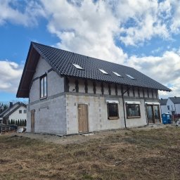 H&R PROJEKT SP. ZO.O. - Surowy dom z betonu komórkowego z czarnym dachem i oknami, w tle inne domy i błękitne niebo z chmurami. Widoczne ślady prac budowlanych.