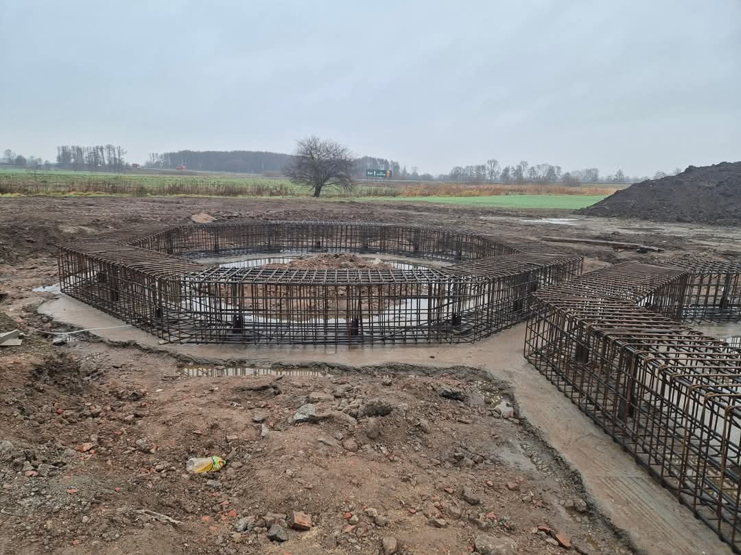Szkielet zbrojenia w kształcie oktagonu na przygotowanym fundamencie pod budowę, widok na rozległe pole i pochmurne niebo.