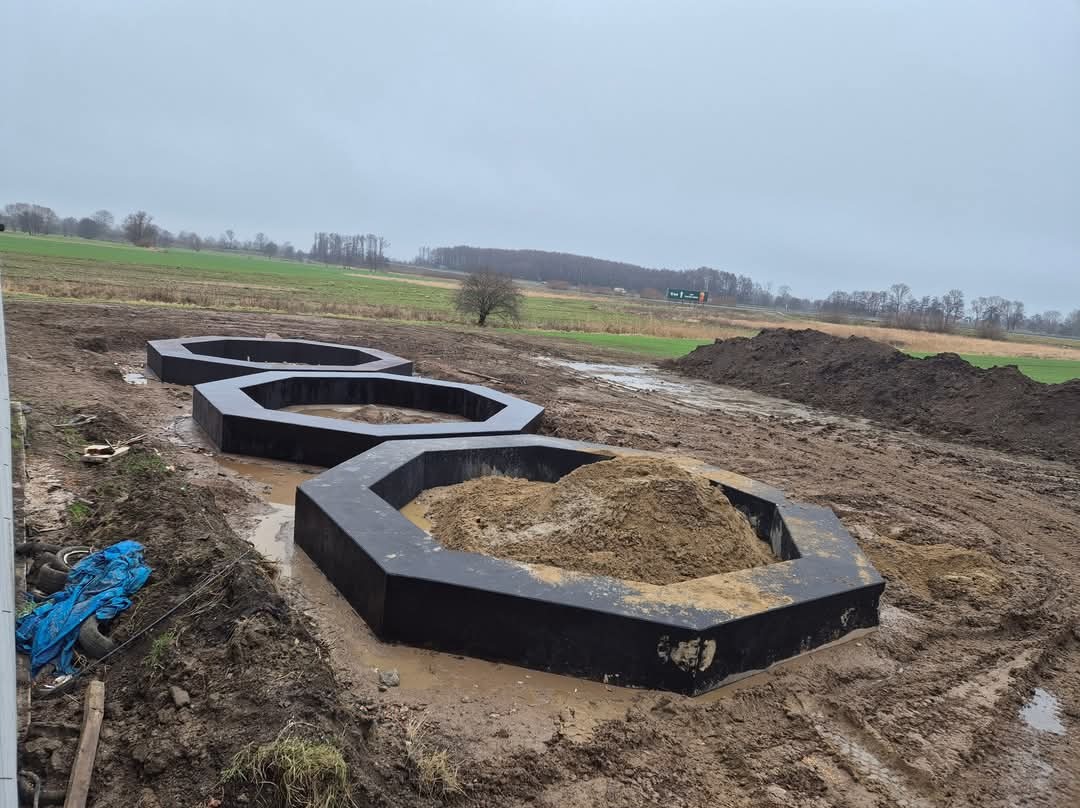 Trzy czarne, betonowe, ośmiokątne obudowy fundamentów w ziemi, wypełnione piaskiem i wodą, na tle pola i pochmurnego nieba.