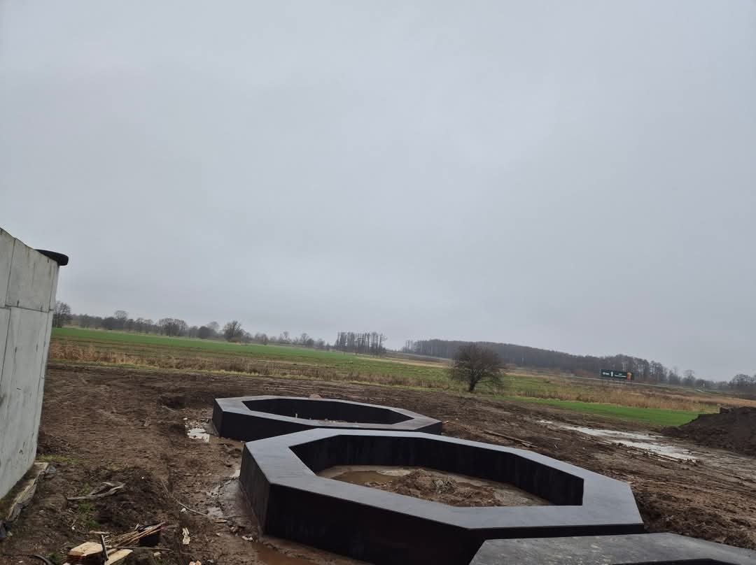Dwa betonowe, heksagonalne pierścienie fundamentowe w grząskim terenie pod budowę, widok na pole w pochmurny dzień.