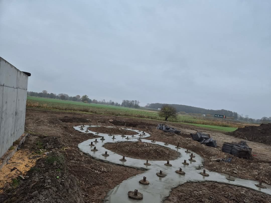 Wykop pod fundamenty z betonowymi stopami, przygotowany pod budowę. Widok na pole i pochmurne niebo w tle. Etap prac ziemnych i betonowania.