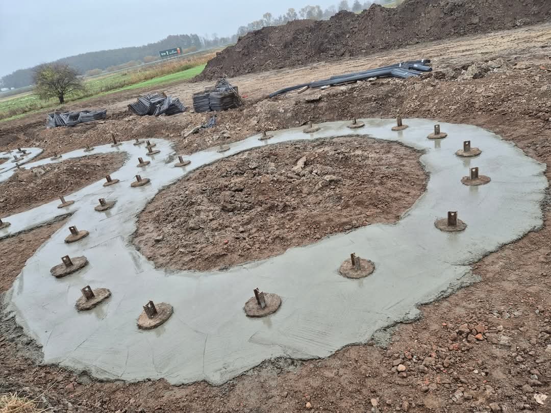 Wylany fundament pod budowę z widocznymi metalowymi elementami mocującymi. Na drugim planie ziemia, trawa i drzewa. Pochmurny dzień.