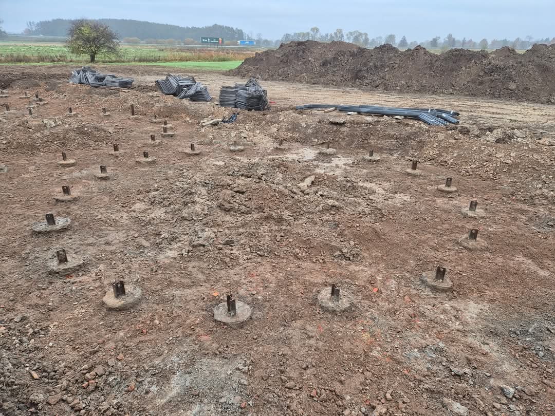 Przygotowany teren pod fundamenty z widocznymi betonowymi słupkami i wystającymi prętami. W tle sterta ziemi i materiały budowlane.