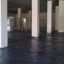 Przygotowanie podłoża: czarna folia izolacyjna rozłożona na podłodze surowego wnętrza z białymi kolumnami, materiały budowlane w tle.