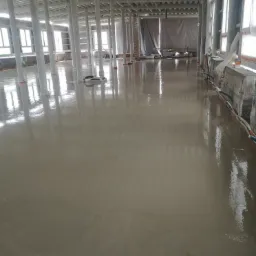 Świeżo wylana, gładka posadzka w surowym stanie, z widocznymi kolumnami i oknami w tle, odbijająca światło w mokrej powierzchni.