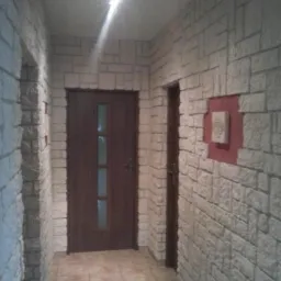 Korytarz z dekoracyjną okładziną imitującą kamień, brązowe drzwi z szybą, płytki podłogowe w odcieniach beżu, oświetlenie sufitowe.