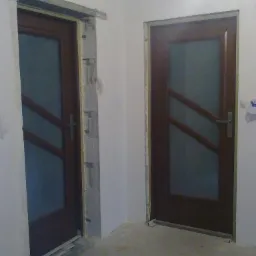 Montaż dwóch brązowych drzwi z szybami w Żninie. Jedne drzwi w trakcie montażu, z widoczną pianką montażową. Drugie drzwi już zamontowane, gotowe do użytku.