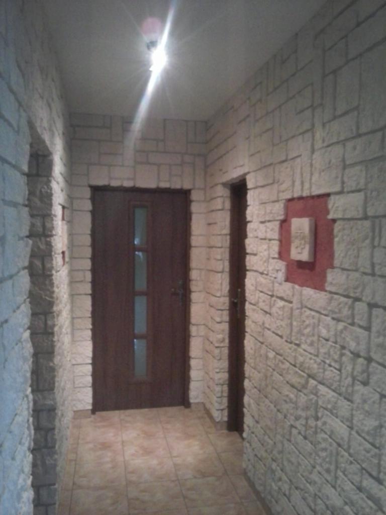 Korytarz z dekoracyjną okładziną imitującą kamień, brązowe drzwi z szybą, płytki podłogowe w odcieniach beżu, oświetlenie sufitowe.