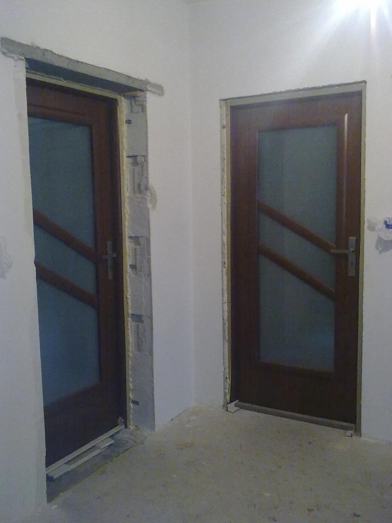 Montaż dwóch brązowych drzwi z szybami w Żninie. Jedne drzwi w trakcie montażu, z widoczną pianką montażową. Drugie drzwi już zamontowane, gotowe do użytku.