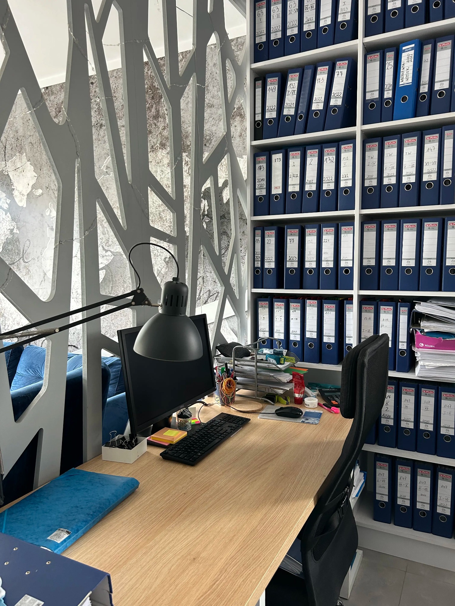 Biuro z biurkiem z jasnego drewna, czarnym monitorem i lampką, regał z segregatorami w tle, element dekoracyjny na ścianie w kształcie drzewa