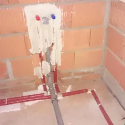 Instalacja wodna w surowym wnętrzu: czerwone rury PEX na podłodze z izolacją, szare rury pionowe w ścianie z cegły ceramicznej, z widocznymi przyłączami wody.