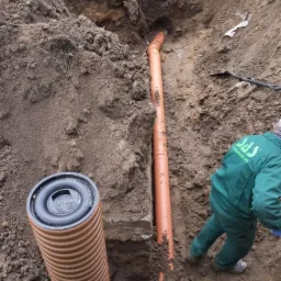 Instalacja rur kanalizacyjnych w wykopie ziemnym, widoczny pracownik w zielonym kombinezonie. Rury z tworzywa sztucznego w kolorze pomarańczowym, studzienka kanalizacyjna.