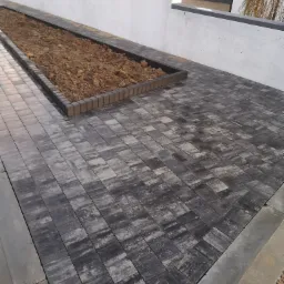 Ułożona szara kostka brukowa z obrzeżem, przylegająca do podwyższonej rabaty z ziemią, widok z perspektywy chodnika.