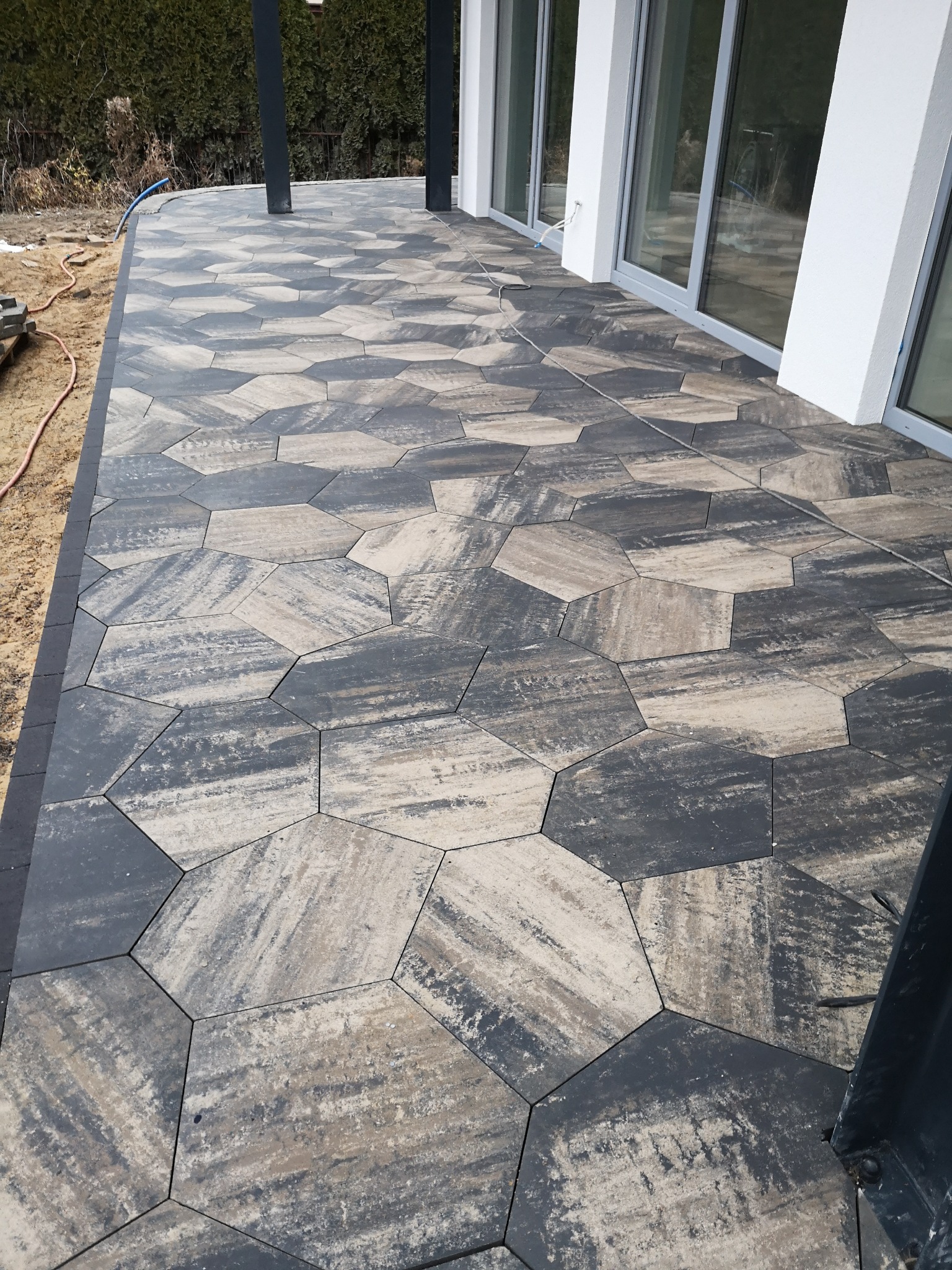 Ułożona nawierzchnia z sześciokątnej kostki w odcieniach szarości i beżu, tworząca wzór na tarasie przylegającym do domu z dużymi oknami przesuwnymi.