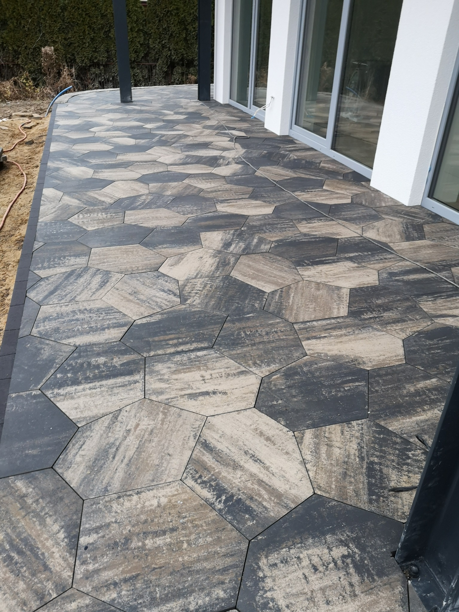 Ułożona sześciokątna kostka w odcieniach szarości i beżu tworzy wzór na tarasie przy nowoczesnym domu z dużymi oknami przesuwnymi.