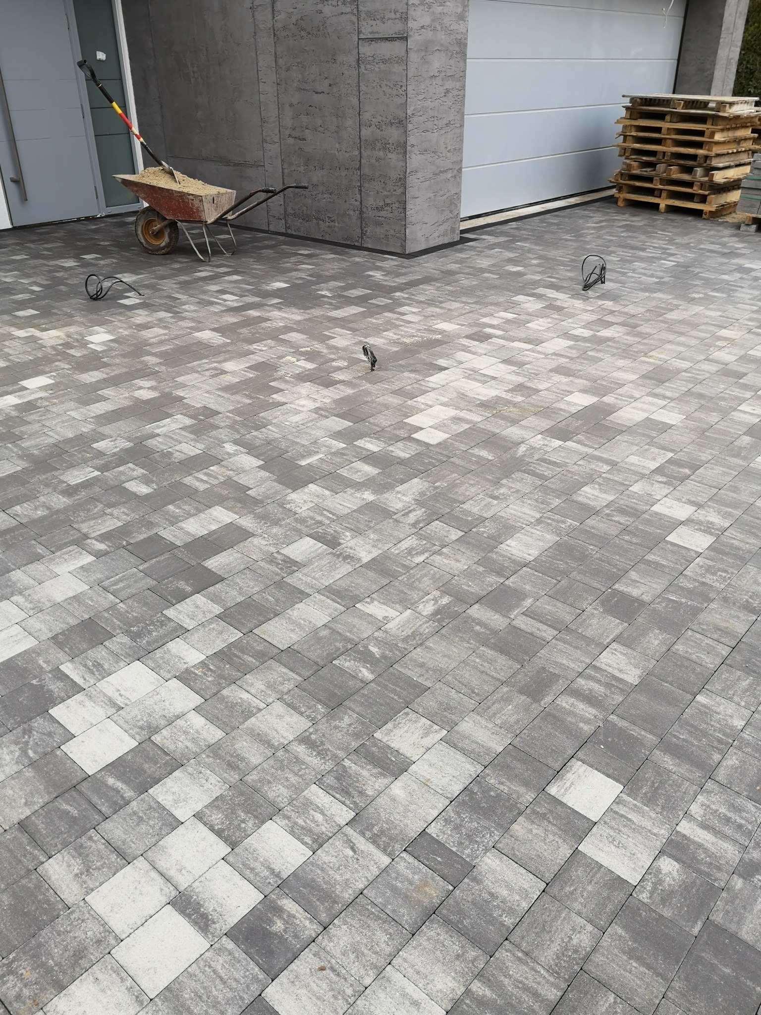 Ułożona szara kostka brukowa przed garażem, taczka z piaskiem i palety w tle.