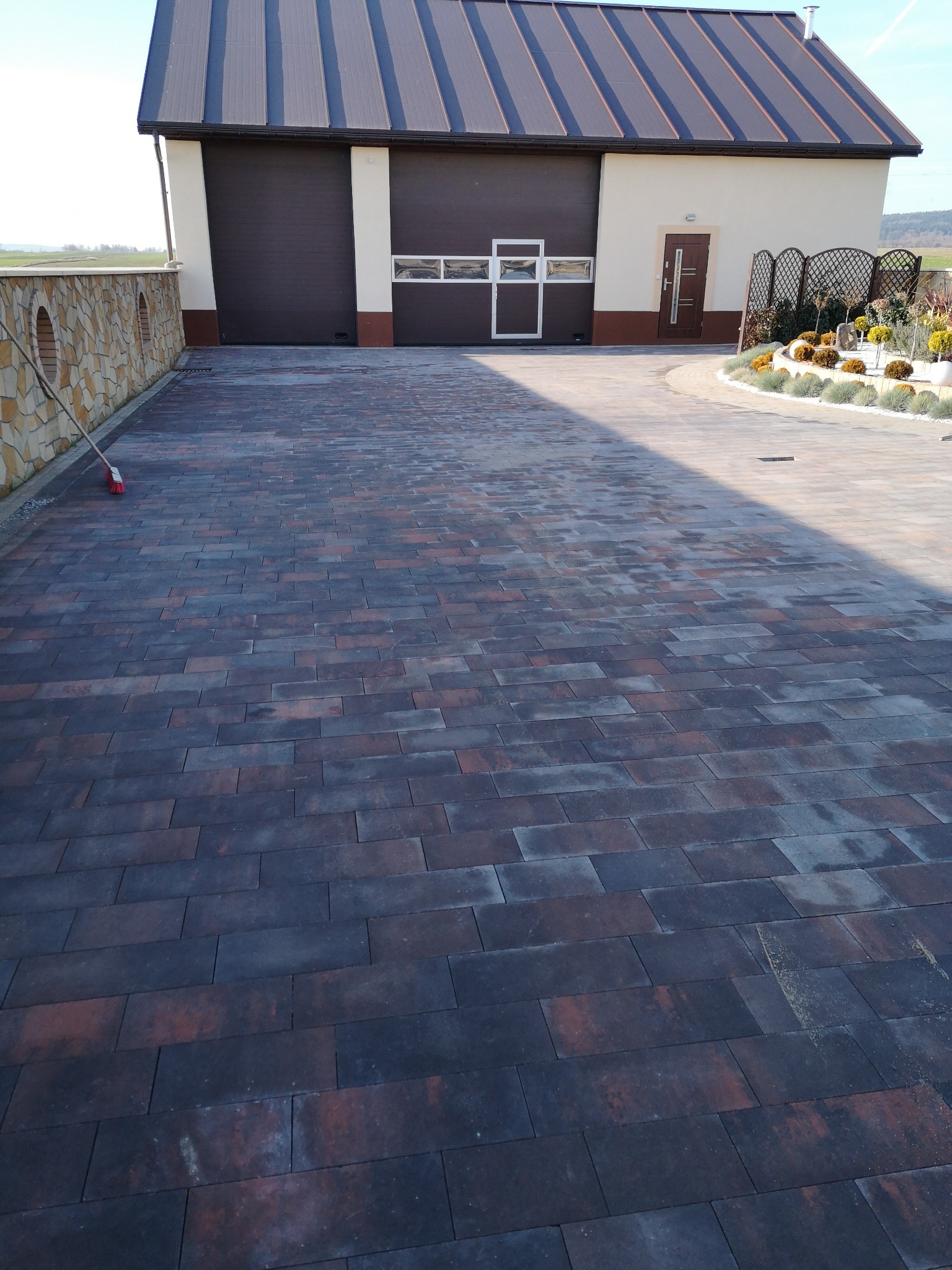 Ułożona kostka brukowa w odcieniach brązu i szarości na podjeździe przed budynkiem z garażem, widoczny fragment kamiennego murku i roślinność ozdobna.