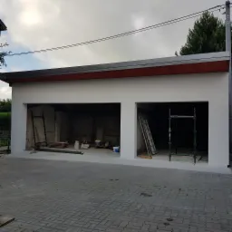 Świeżo otynkowany garaż dwustanowiskowy z czerwonym podbitką, widoczne rusztowania i drabiny wewnątrz, kostka brukowa przed budynkiem.