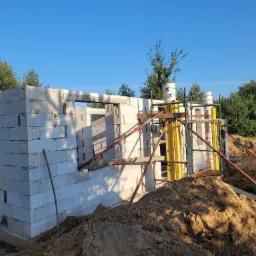 Wznoszenie ścian domu z bloczków betonu komórkowego, widoczne podpory szalunkowe i hałda ziemi na pierwszym planie, w tle drzewa i błękitne niebo.