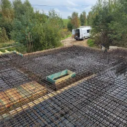 Zbrojenie płyty fundamentowej z widocznym szalunkiem i stojącym w tle samochodem dostawczym na tle zieleni.
