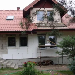 Dom jednorodzinny w trakcie prac elewacyjnych: ocieplanie styropianem, częściowo wykończony dach z oknem, rusztowanie i drewno opałowe przed budynkiem.