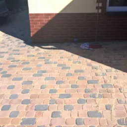 Ułożona kostka brukowa w odcieniach czerwieni i szarości, z widocznym fragmentem elewacji budynku i samochodu. Na kostce leżą narzędzia.