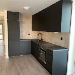 Nowoczesna kuchnia z zabudową w kolorze grafitowym, minimalistyczne fronty bez uchwytów, grafitowa armatura, płytki imitujące beton na ścianie, jasna podłoga drewnopodobna.