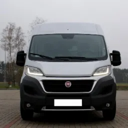 Przód białego furgonu Fiat Ducato na brukowanym placu, światła włączone, widoczne drzewa w tle, pochmurna pogoda.