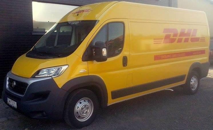 Żółty samochód dostawczy DHL na zewnątrz budynku, widok z boku, oświetlenie dzienne. Auto z widocznym logo firmy.