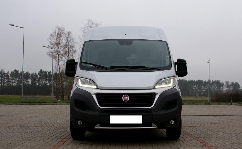 Przód białego furgonu Fiat Ducato na brukowanym placu, światła włączone, widoczne drzewa w tle, pochmurna pogoda.