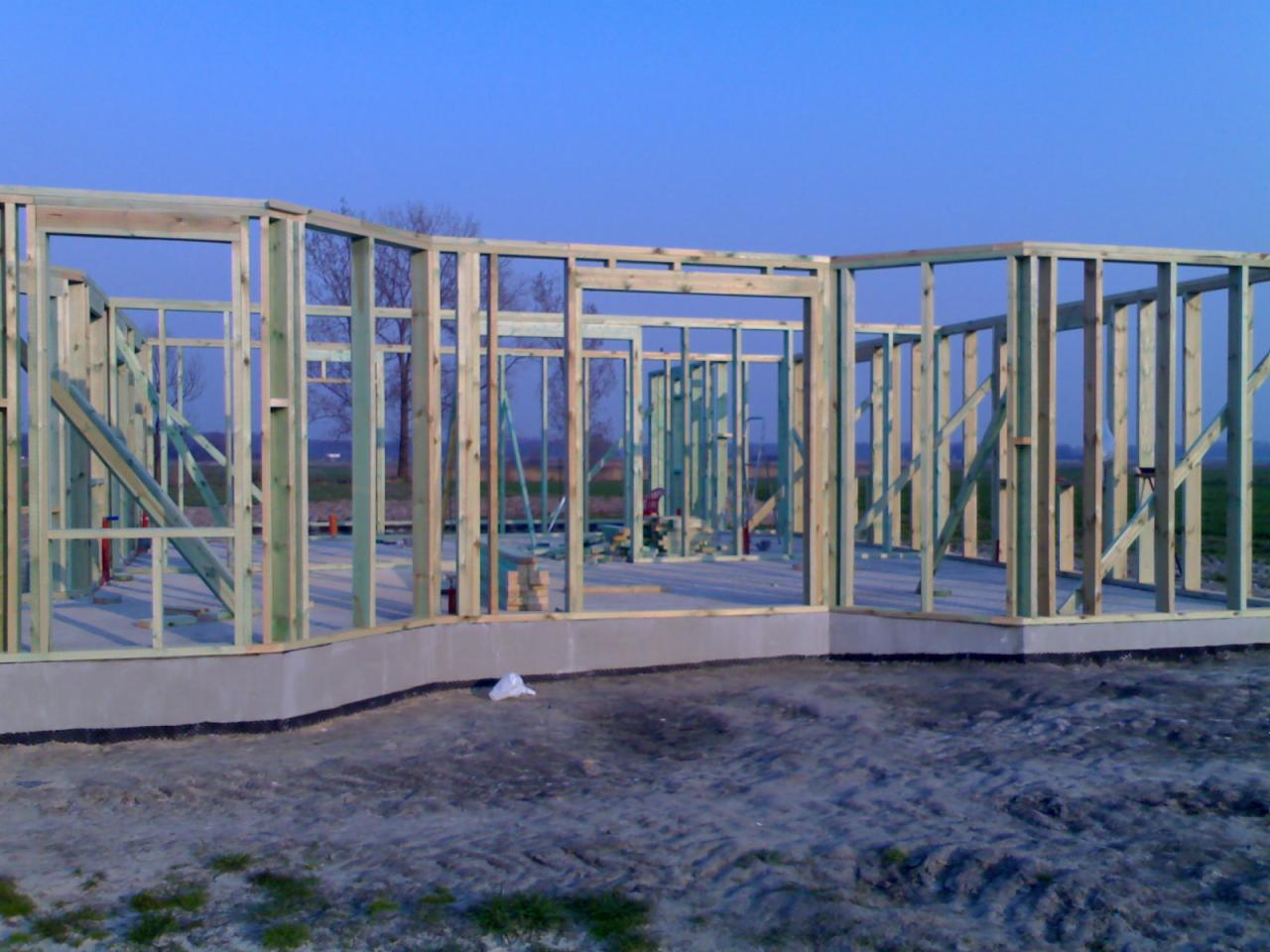 Wznoszenie konstrukcji szkieletowej domu na fundamencie. Widoczne drewniane elementy ścian i otworów okiennych. W tle błękitne niebo i drzewa.